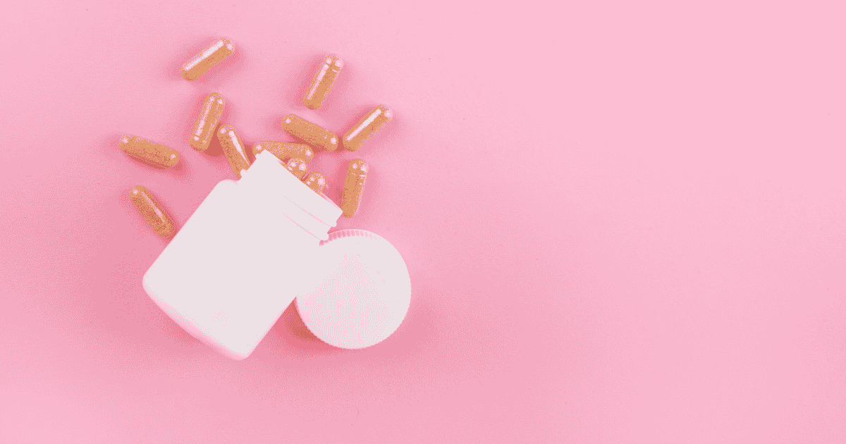 An open white supplement bottle spills light brown capsules onto a solid pink background.