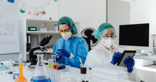 Two laboratory researchers wearing protective gear and masks analyzing data and handling scientific equipment.