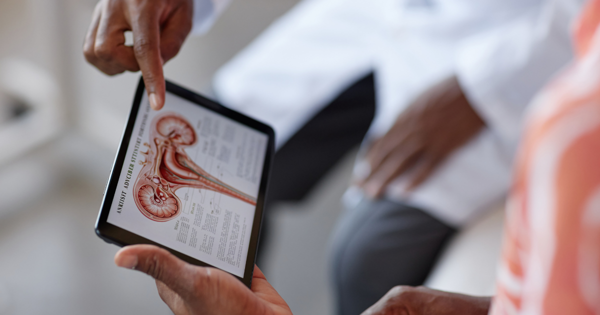 Over-the-shoulder view of a doctor pointing to an anatomical medical diagram displayed on a tablet screen during a consultation.