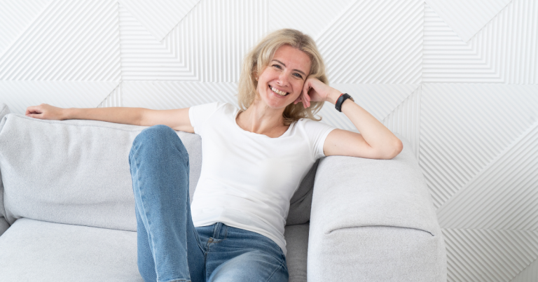 A smiling woman with blonde hair wearing a white t-shirt and jeans relaxing on a light grey couch.