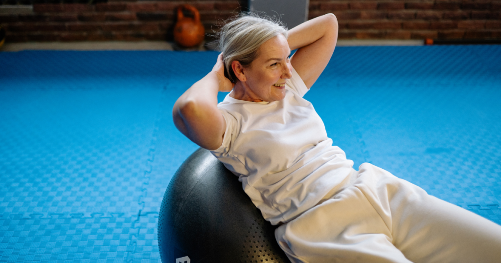 A smiling older woman in white exercise clothes leaning back on a dark fitness ball on a blue mat.